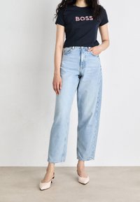 Granatowa koszulka z napisem "BOSS" w kwiatowym nadruku, połączona z jasnoniebieskimi spodniami typu straight-leg i jasnoróżowymi kobiecymi klapkami na obcasach. Standardowy krój, casualowy styl.