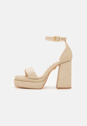 Arany csillogós platform szandál széles elülső pánttal, bokapánttal és vastag sarokkal. Fém csattal és sima talppal rendelkezik.