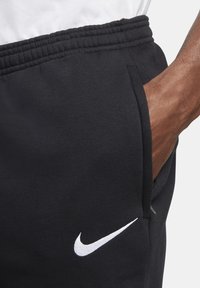 Närbild av en persons hand i fickan på svarta Nike sweatpants med en vit logotyp och elastisk midja.