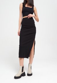 Czarny crop top i dopasowana czarna midi spódnica z bocznym rozcięciem. Model nosi beżowe botki za kostkę z grubą czarną podeszwą. Gładka faktura materiału.