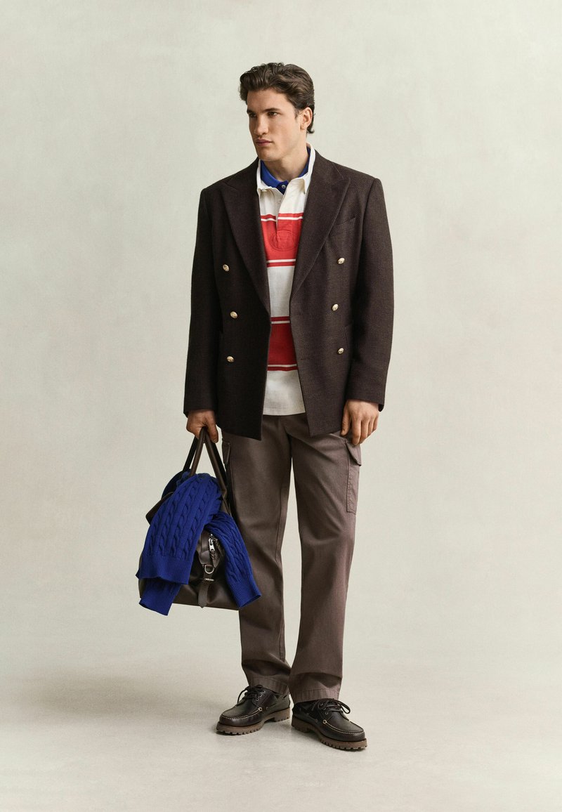 Mann trägt braunen zweireihigen Blazer, gestreiftes Poloshirt, braune Hose, schwarze Schuhe, hält schwarze Tasche, über die ein blauer Pullover gelegt ist.