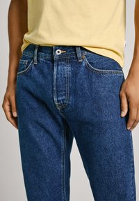 Jeans de denim azul con corte recto, que cuentan con cierre de botón, costuras contrastantes y cinco bolsillos. Llevados con una camisa amarilla.