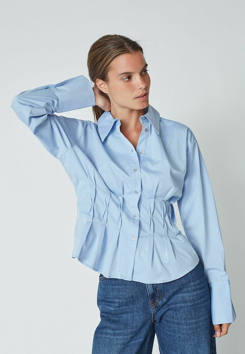 Light blue button-up shirt with long sleeves, featuring gathered texture at the waist and a classic collar. Paired with blue jeans.