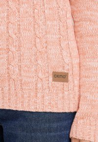 Lys rosa kabelstrikket genser med ribbestruktur og tettsittende ermer. Har et brunt skinnlapp med logoen "OXMO" på siden.