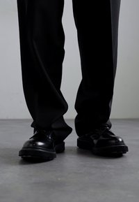 Zwarte leren schoenen met een ronde neus, gestructureerde rubberen zool en vetersluiting, gecombineerd met losse zwarte broeken. Achtergrond van een betonnen vloer.