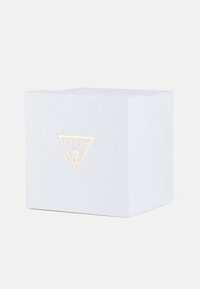 Biała teksturowana pudełko z złotym trójkątnym logo z napisem "GUESS". Pudełko ma czyste linie i minimalistyczny design.