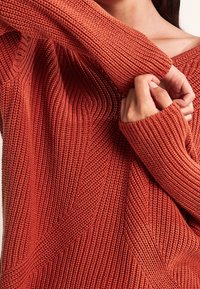 Sweter w kolorze rdzawego brązu, wykonany na drutach, z teksturowanym wzorem, z pionowymi i ukośnymi motywami oraz szerokimi ściągaczami.