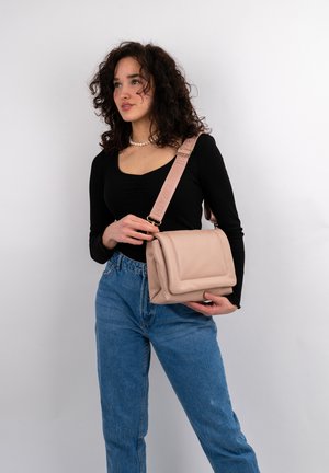 Borsa a tracolla in pelle rosa chiaro con un design strutturato, piega decorativa e una strap larga con logo in rilievo. Il modello la tiene in modo informale.