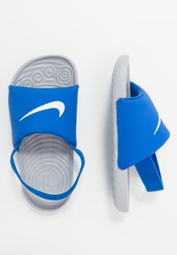 Sandale bleue avec une large lanière et un logo swoosh blanc. Semelle texturée grise, rembourrée pour un meilleur confort, et un entre-doigts en caoutchouc pour un maintien sécurisé.