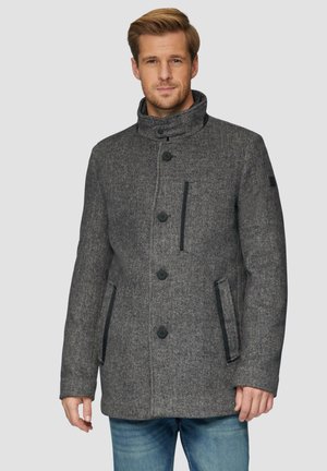 Manteau en chevron gris avec un col montant, doté de boutons noirs et de poches latérales, porté par-dessus une chemise bleu clair et un denim.
