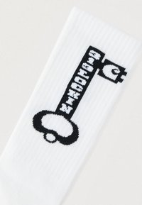 Λευκή κάλτσα με μαύρο γραφικό ενός κλειδιού και στυλιζαρισμένο κείμενο ενσωματωμένο στο σχέδιο του κλειδιού.