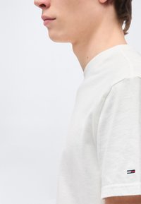 Νεαρό άτομο που φοράει ένα απλό λευκό κοντομάνικο μπλουζάκι με ένα μικρό κόκκινο, άσπρο και μπλε λογότυπο στο μανίκι, κοιτάζοντας πλάγια.