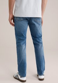 Jeans de mezclilla azul con corte recto, detalles desgastados y bolsillos traseros. Combinados con zapatillas blancas con rayas negras.