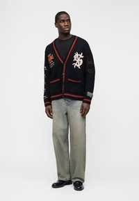 Uomo in piedi che indossa un cardigan nero con bordi rossi e toppe, maglietta nera, jeans grigi larghi e scarpe nere, che guarda leggermente di lato.