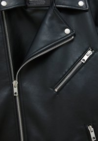 Veste de moto en cuir noir avec fermetures éclair et boutons-pression argentés, affichant les détails du col et de la poche poitrine zippée.