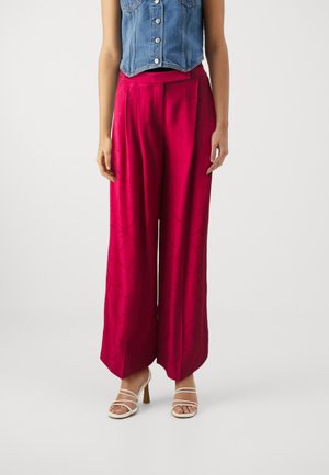 Loose-fitting, high-waisted red trousers with a smooth texture and subtle circular pattern. Paired with light-colored strappy heeled sandals.