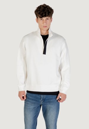 Pullover bianco con zip a metà e accento nero, con polsini e vita a coste. Indossato sopra una maglietta nera, abbinato a jeans blu. Tessuto dalla texture liscia.