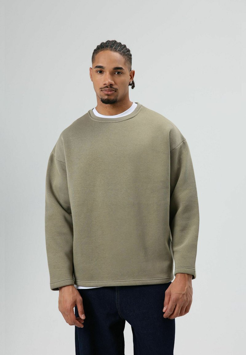 Oudgroene oversized sweater met lange mouwen, ronde hals, zachte textuur en zichtbare stiksels. Gedragen over een wit T-shirt en gecombineerd met donkere jeans.