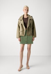 Giacca in ecopelle verde oliva chiaro con ampio colletto, accessori in metallo argentato e apertura frontale. Abbinata a una gonna in corduroy verde e sandali con tacco trasparente.