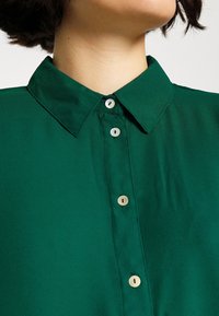 Groen overhemd met knopen en een kraag. Gemaakt van gladde stof, met vijf kleine, ronde, lichtgekleurde knopen aan de voorkant.