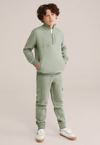 Šviesiai žalias sportinis kostiumas, kuriame yra zip-up puloveris su kengūros kišenėmis ir derančios jogger kelnes su šoninėmis kargo kišenėmis ir raukinėtais rankogaliais.