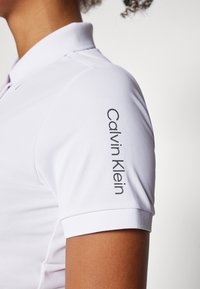 Balta polo marškinėliai su klasikiniu apykaklės dizainu, pasižymintys tekstūruota medžiaga ir akivaizdžiu Calvin Klein logotipu, spausdintu ant rankovės.