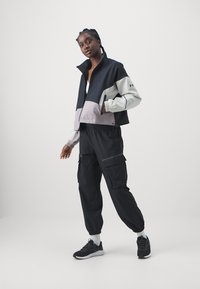 Schwarze und graue Sportjacke mit einem kurzen Design, hohem Kragen und Reißverschluss. Schwarze Cargo-Hosen mit elastischen Bündchen. Schwarze Sneakers.