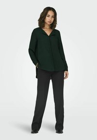 Camicia button-up verde scuro con scollatura a V, maniche lunghe e una leggera lucentezza, abbinata a pantaloni neri a gamba larga e stivaletti con tacco.