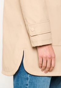 Beżowa wodoodporna kurtka z rękawem i detalem z guzikiem, z gładką fakturą i bocznym wentylkiem. Noszona na odzieży dżinsowej.