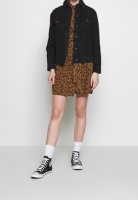 Černá džínová bunda s předními knoflíky, přes hnědé šaty s černým leopardím vzorem. Bílá ponožky a černé vysoké tenisky dotvářejí celkový outfit.