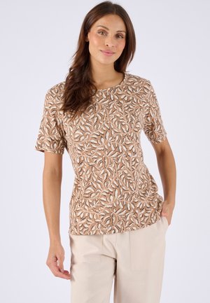 Frau trägt ein beiges kurzärmeliges Oberteil mit einem weißen und braunen Blattmuster und helle Hosen vor einem einfarbigen Hintergrund.