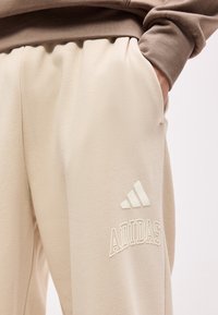 Μπεζ παντελόνι φόρμας Adidas με λευκό λογότυπο στον μηρό, φορεμένο με καφέ μπλούζα μακρυμάνικη. Χέρι ορατό στην τσέπη του παντελονιού.