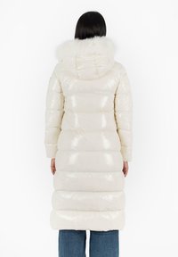 Lungo cappotto imbottito in tessuto crema lucido, con cappuccio foderato in pelliccia, polsini a coste e sezioni orizzontali trapuntate per l'isolamento.