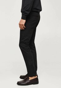 Schwarze, schmal geschnittene Hosen aus Baumwolle mit glatter Textur und klassischem Design. Kombiniert mit dunkelbraunen Leder-Loafers.