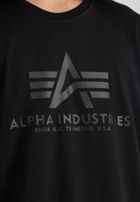 Crna pamučna majica s istaknutim sivim grafičkim logotipom sa dva pruga i tekstom "ALPHA INDUSTRIES, KNOXVILLE, TENNESSEE, U.S.A."
