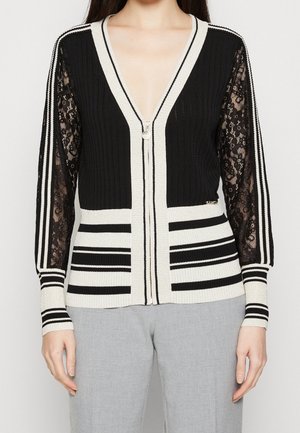 Femme portant un cardigan zippé noir et blanc avec des manches en dentelle et un motif rayé, associé à un pantalon gris clair.