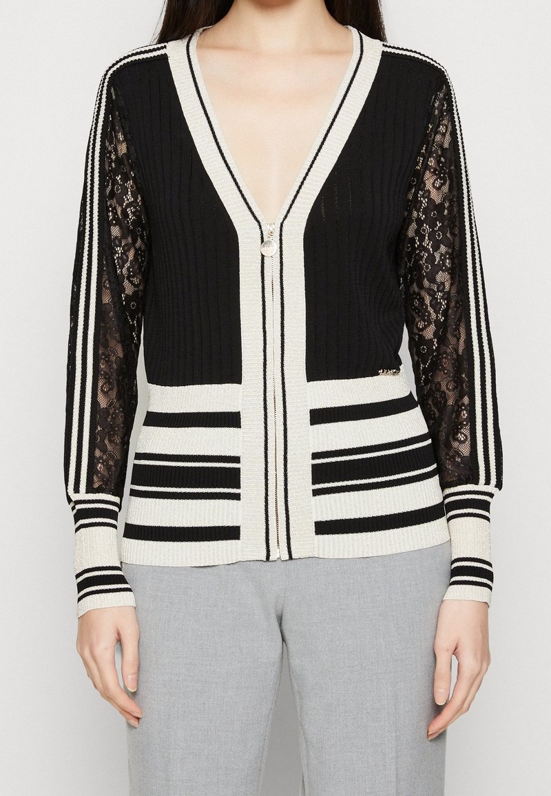 Femme portant un cardigan zippé noir et blanc avec des manches en dentelle et un motif rayé, associé à un pantalon gris clair.