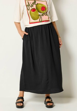 Kvinde iført hvid T-shirt med grønne olivenprint og sort maxinederdel med lommer, kombineret med sorte plat-sandaler.