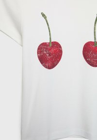 Vit bomullströja med två röda körsbärstryck, var och en med gröna stjälkar. Designen har en något slitagen textur.