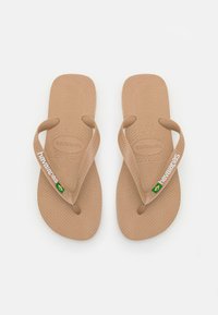 Tan spalvoti guminiai šlepetės su tekstūruotais pado paviršiais. Plonos dirželiai su baltu „Havaianas“ ženklinimu ir mažu Braziliškos vėliavos akcentu.