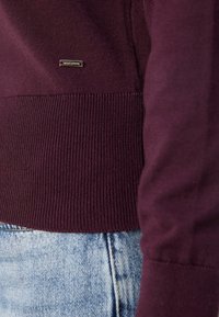 Gros plan sur l'ourlet et la manche d'un pull en maille bordeaux associé à un jean denim bleu clair, avec une petite étiquette de marque métallique rectangulaire.