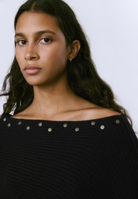 Suéter negro de punto con escote off-shoulder y acentos de tachuelas metálicas a lo largo del cuello. Textura suave y corte relajado.