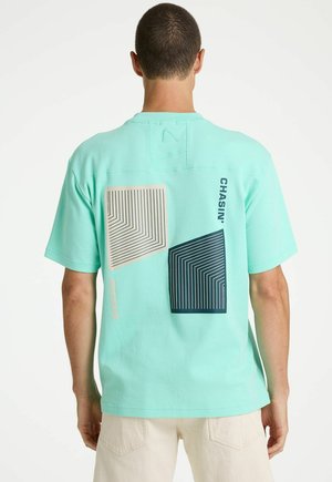 Man draagt een mintgroen T-shirt met twee geometrische vierkante ontwerpen en de tekst "CHASIN'" op de bovenrug, gecombineerd met lichtbeige broek.