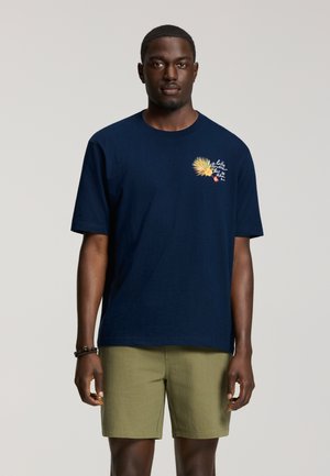 Uomo che indossa una maglietta blu navy con grafica gialla di foglia di palma e testo sul petto, abbinata a pantaloncini verde oliva, in piedi davanti a uno sfondo grigio.