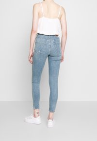 Jeans skinny de tiro alto en azul claro con cinco bolsillos, ligero desgaste y una textura envejecida, combinados con una camiseta sin mangas blanca y zapatillas blancas.