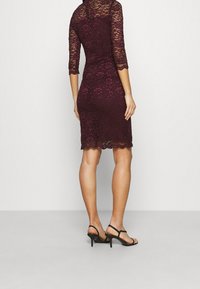 Robe en dentelle bourgogne à manches trois-quarts et ourlet festonné, associée à des sandales à talons hauts noires, mettant en valeur une silhouette ajustée.