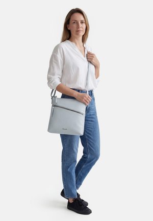 Mujer con blusa blanca, vaqueros azules y zapatos negros, llevando un bolso cruzado gris claro con bolsillo frontal con cremallera.