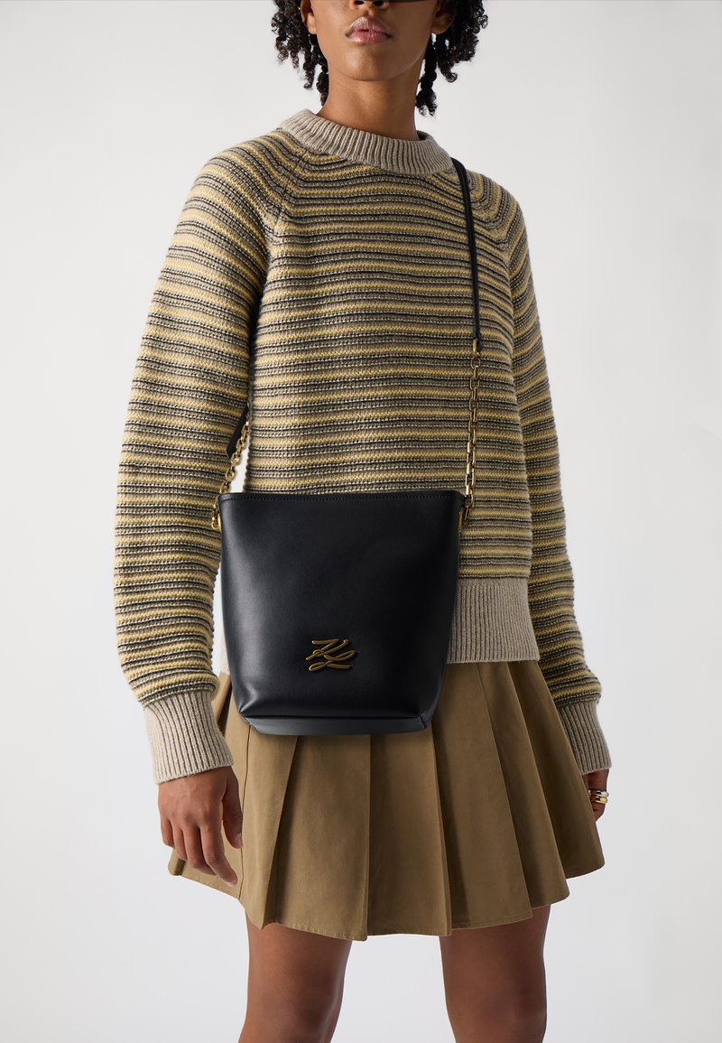 Černá kožená crossbody taška se zlatými detaily, vyznačující se hladkým povrchem a jednoduchým siluetou. Spárována s pruhovaným svetrem a skládací sukní.