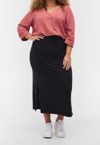 Blouse rose, boutonnée, à manches trois-quarts, associée à une jupe noire, ajustée, mi-longue avec une fente sur le côté. Baskets blanches.