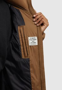 Hnědý prošívaný kabát s vycpávkou, s černým interiérem a štítkem s nápisem: "Teplé objetí vyrobené z látky."